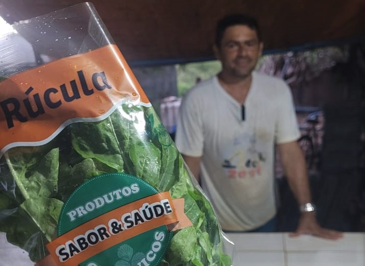 Cedida Pacote de rúcula aparece em primeiro plano com o produtor Tiago ao fundo de camisa branca